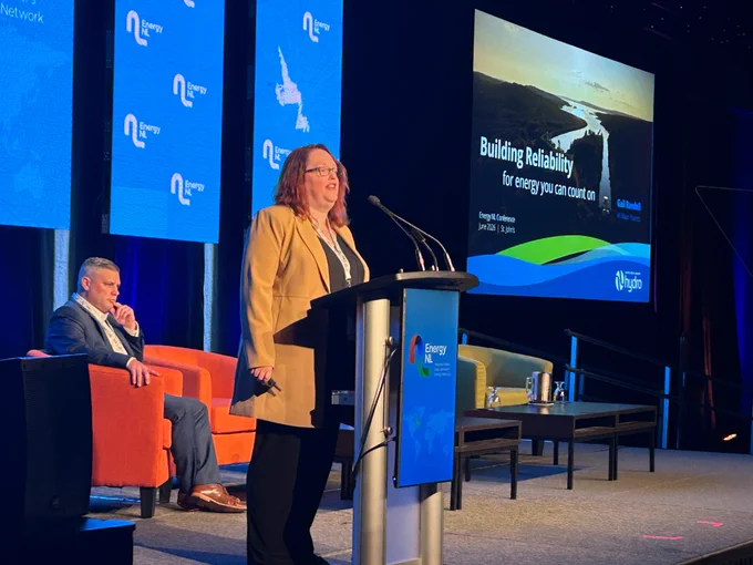 Hydro VP Gail Randell speaks from a podium on the conference stage, with a presentation on screen in the background. A man sits on stage in a chair near her, listening. 