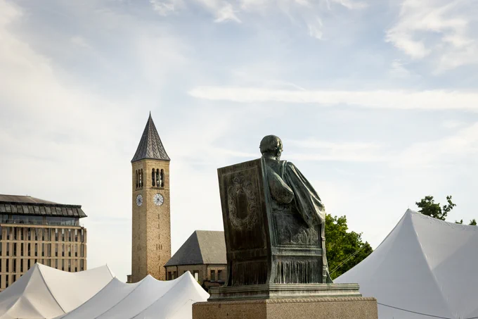 The AD White statue on the Cornell University Arts Quad sits in the foreground with its back facing the viewer, overlooking a scene with white tents at the 2026 Cornell Reunion. The McGraw Tower at Cornell University is visible in the background, under a partly cloudy sky.
