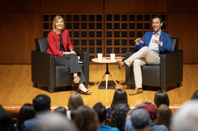 Two people sitting and smiling in armchairs on a stage, engaged in a conversation. A small round table with cups is between them. There is an attentive audience in the foreground.