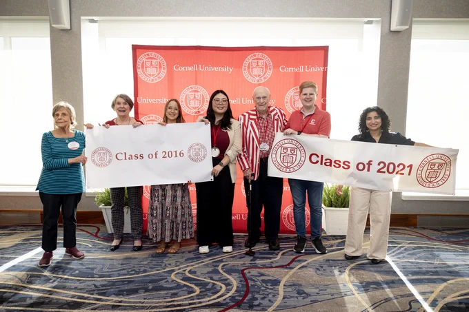 A group of people stand indoors, holding banners that read "Class of 2016" and "Class of 2021" in front of a red Cornell University backdrop. They are smiling, and some are wearing casual attire while others have more formal clothing.