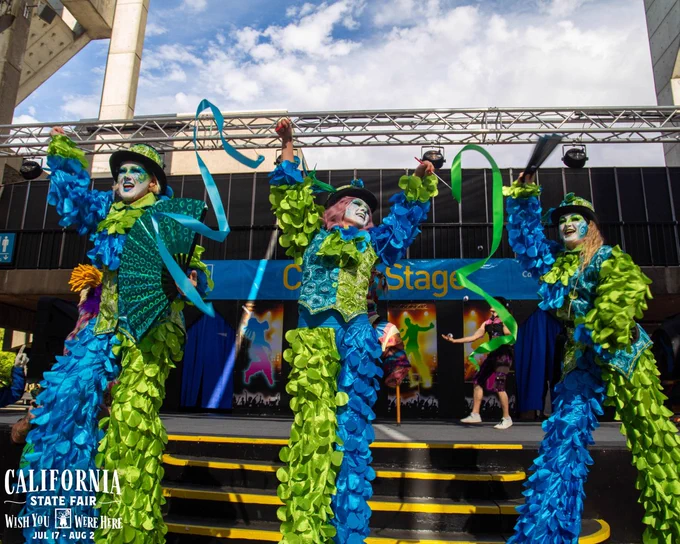 Performers in vibrant blue and green costumes dance energetically on a stage during Poppy's Birthday at the California State Fair.
