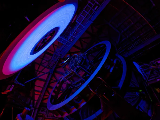 An ominous blue donut-shaped ring surrounded by a thin red ring glows ominously in the dark to the left. To the right, Rubin's large steel telescope mount structure is barely visible, softly illuminated by the glow.