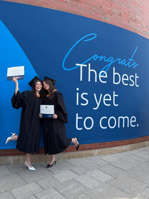 A photo of two graduates posing in front of a sign that says “Congrats, the best is yet to come.”
