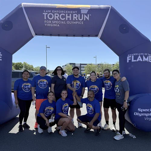 Group of runners wearing matching blue Law Enforcement Torch Run shirts posing under the event's inflatable arch outdoors.