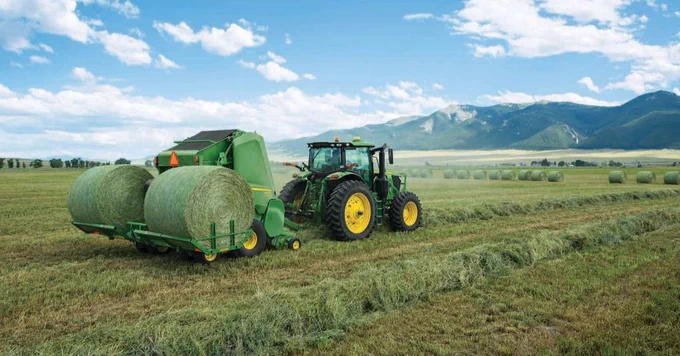 A green tractor is towing a hay baler in a large field, producing round bales of hay. The landscape features open farmland with mountains in the background under a partly cloudy sky.