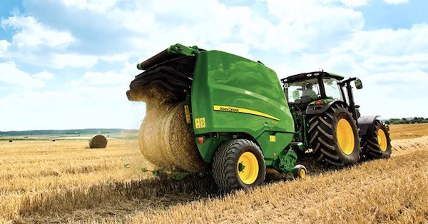 A John Deere tractor operates in a large field, baling hay under a partly cloudy sky.