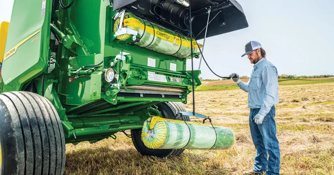 A person stands near a large John Deere agricultural machine in a field. They appear to be inspecting or operating the machinery, which is equipped with hay bales. The grass around is dry and golden under a clear sky.