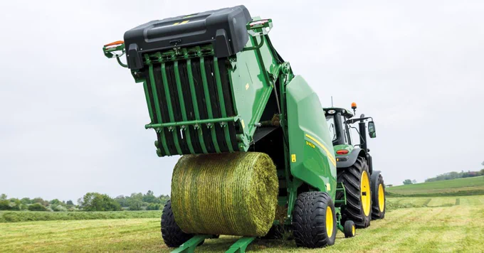 A John Deere tractor is operating in a field, using a baler to produce a large, round hay bale. The machine is vibrant green with yellow accents and is releasing the bale onto the grassy field. The landscape in the background includes lush green grass and a cloudy sky.