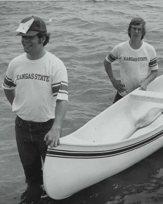 Two K-State students stand in shallow water beside a canoe, in an archival photo from K-State.