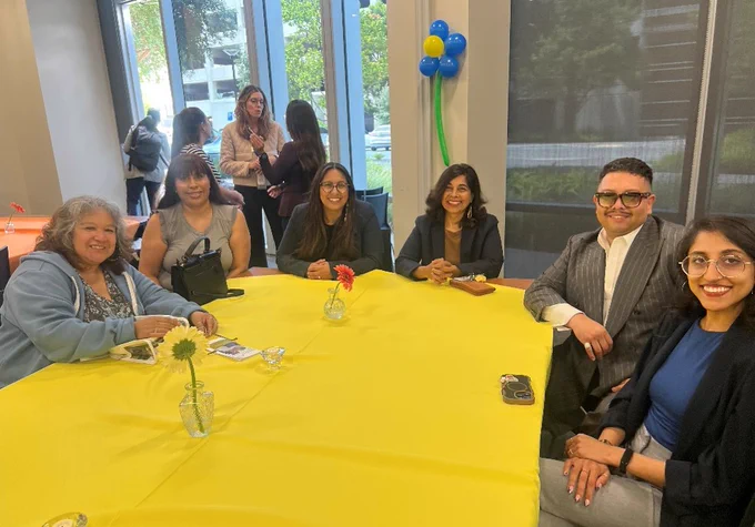 Six attendees sit around a table with a yellow tablecloth, smiling toward the camera during the event.
