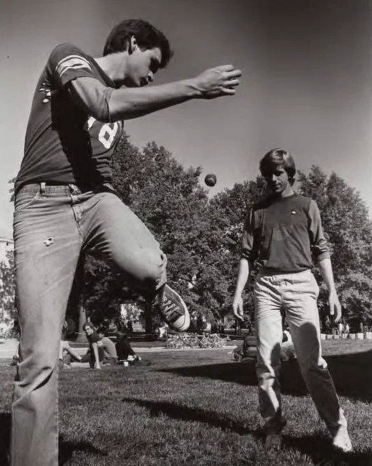 Two individuals playing hacky sack in a grassy area with trees in the background. 