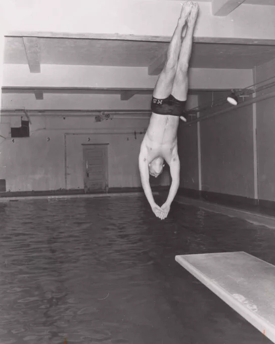 A person dives into an indoor swimming pool, captured mid-air above the water. The diver is wearing a swimsuit with "KS" printed on it. The background shows the pool's interior with a diving board in the foreground.