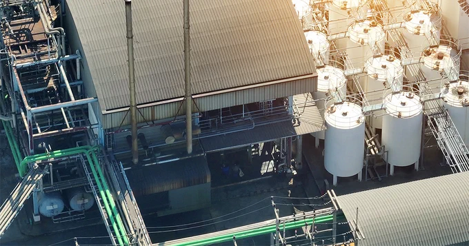 Aerial view of an industrial facility with large storage tanks, pipes, and metal structures. Sunlight illuminates parts of the complex, highlighting the tanks and a corrugated metal roof.