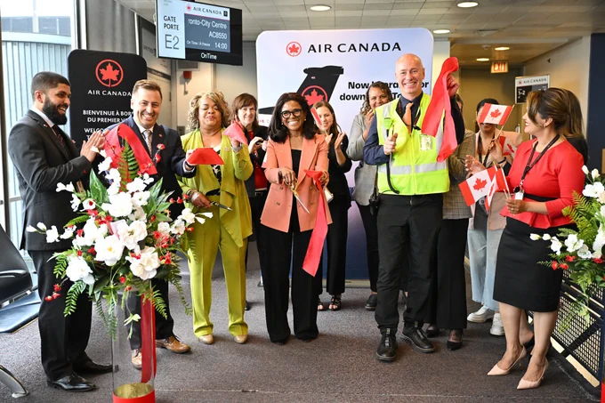 A group of people standing together at an airport event with a ribbon-cutting ceremony. Some individuals are holding Canadian flags and scissors. There are Air Canada logos and signage in the background.