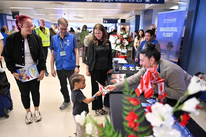 A child is handed a Canadian flag by a person at a table covered with Canadian flags and decorations. Surrounding the child are a few adults, smiling as they observe the interaction. In the background, there are signs for O'Hare Airport and "Inaugural Flight." 