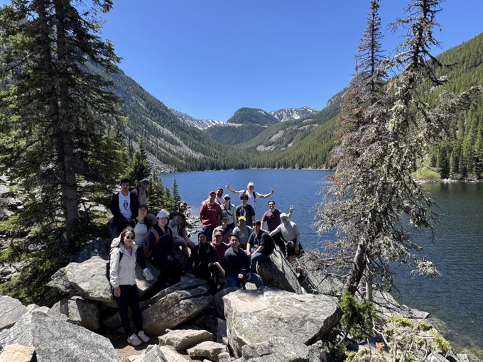 Full-color image of a group standing among the trees next to a beautiful blue lake.