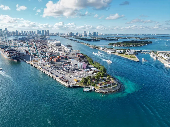 Aerial shot of Miami port with cargo ships, containers, and cranes on a clear, sunny day. Boats sail on blue waters towards islands. City skyline visible in background.