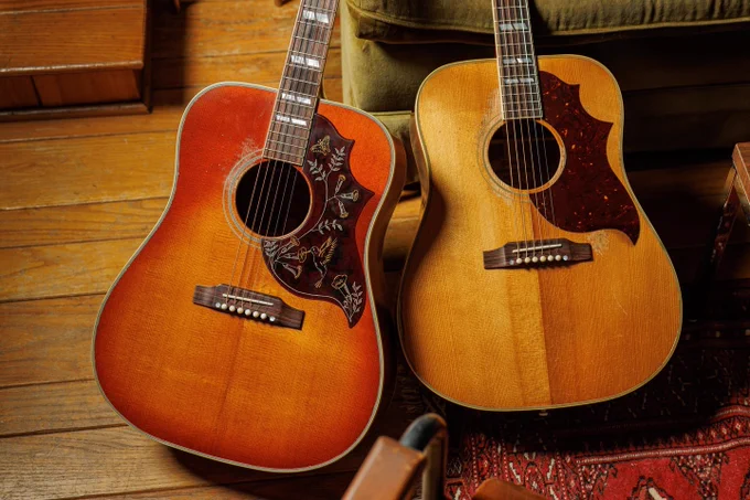 Two acoustic Gibson guitars with decorative pickguards are placed side by side on a wooden floor, near a green sofa.