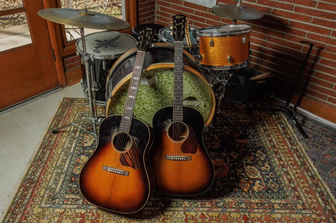 Two sunburst acoustic Gibson guitars are positioned in front of a drum set. They're resting on a patterned rug in a room with brick walls and a wooden door.