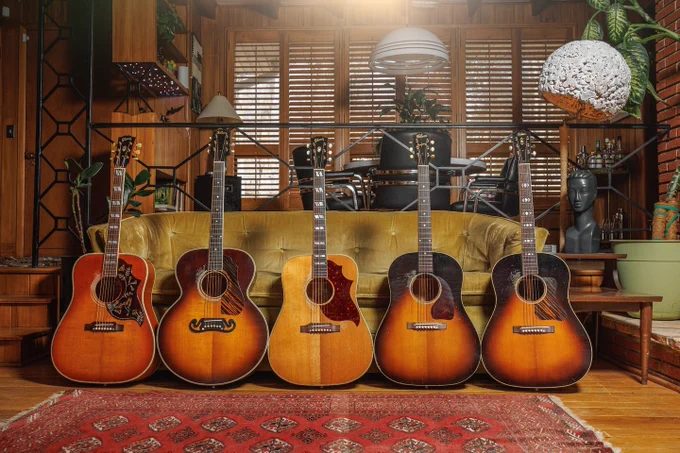 A row of five vintage Gibson acoustic guitars is displayed in front of a yellow couch in a warmly lit room with wooden walls and floors. The guitars vary slightly in design, and a decorative rug is on the floor. Houseplants and unique sculptures are in the background.