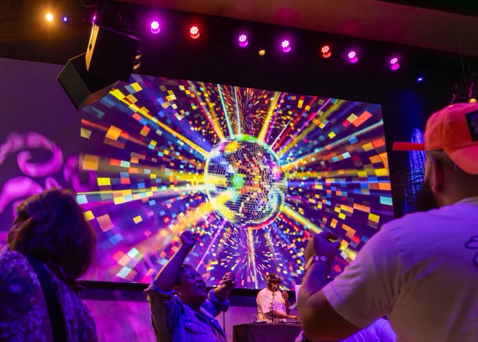 A lively scene featuring a DJ on stage, illuminated by vibrant multicolored lighting. On a large screen behind the DJ, a dazzling digital image of a disco ball with radiating colorful patterns is displayed. People in the foreground are dancing, creating an energetic atmosphere.
