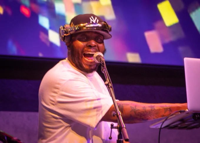 A DJ enthusiastically performing on stage, wearing headphones and a New York Yankees cap, using a microphone and laptop. The background features colorful, abstract light patterns.