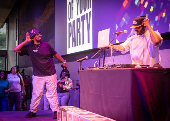A lively DJ performance is taking place on stage. A DJ is standing at a booth with equipment, wearing a cap with "NY" on it, and adjusting headphones. Another performer is on stage, gesturing energetically toward the crowd. 