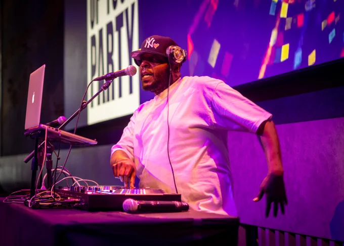 A person in a white shirt and black cap with a "NY" logo is DJing at a setup with a laptop and turntables. 