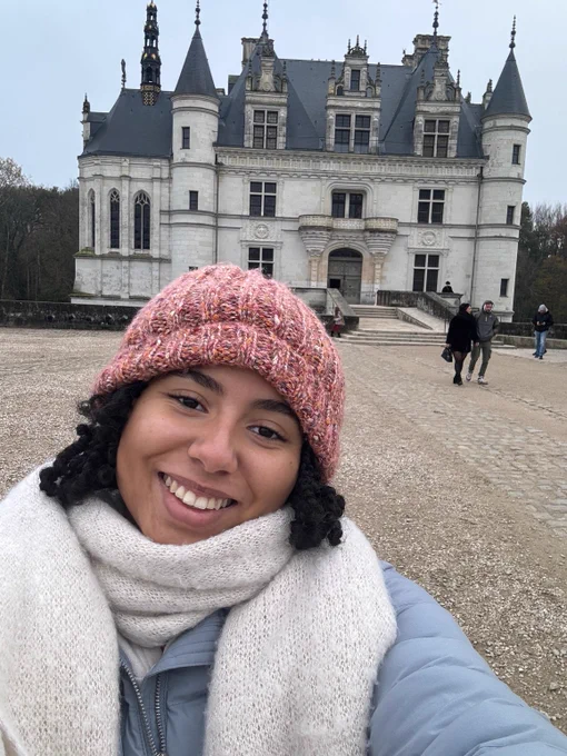 Elise takes a selfie with a scarf and other winter clothing outside in front of a building.