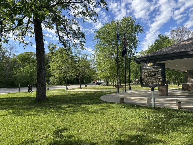 The exterior of the Heath Creek Northbound Rest Area on a sunny day, featuring a green lawn, large trees and flags displayed next to a building with a sloped roof.
