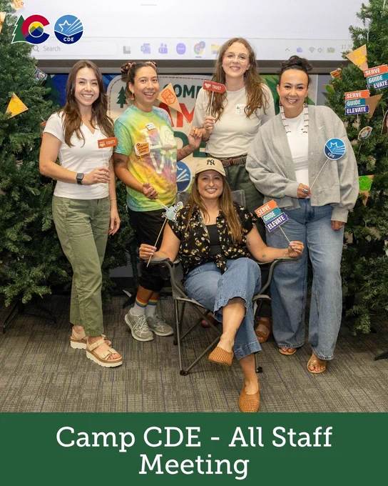 Group photo of five people smiling and standing around a person seated in a camping chair. They are surrounded by artificial pine trees. Some hold small signs with positive, motivational messages. Logos for Google Search and CDE appear at the top. Text at the bottom reads, "Camp CDE - All Staff Meeting.