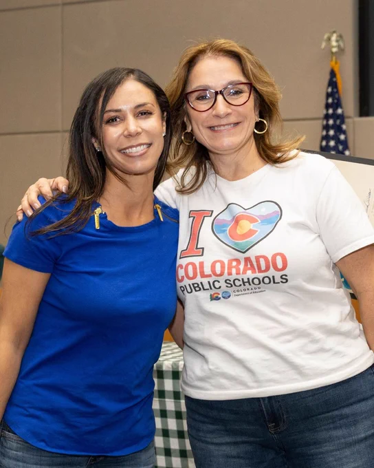Two people smiling and posing together indoors. One person is wearing a blue shirt, and the other is wearing a white "I Love Colorado Public Schools" T-shirt with the Colorado flag emblem. An American flag is visible in the background.