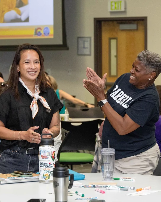 Two people are engaged in a lively interaction in a classroom setting. One individual wearing a scarf is smiling and standing near a table with notebooks and water bottles. Another individual, in a T-shirt that says "Farrell," is clapping and smiling. In the background, a presentation screen and a door are visible.