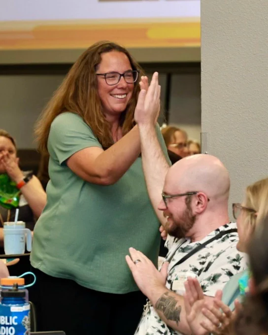 One person stands while another sits in a crowded room. They high-five each other as the standing person walks past.