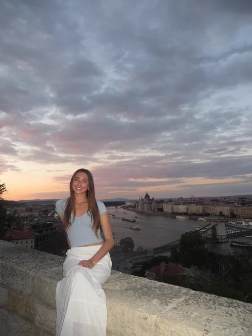 Person sitting on a stone ledge with a scenic view of the Danube River. The background includes the cityscape of Budapest with notable landmarks like the Hungarian Parliament Building under a partly cloudy sky at sunset.