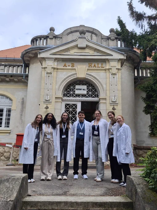 A group of people wearing white lab coats stand in front of an ornate building labeled "A.B Hall." They are smiling and have name tags, posed on steps leading up to the entrance. The architecture features columns and decorative elements.