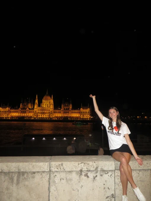 A person sitting on a stone ledge at night, smiling and raising one arm. The illuminated Hungarian Parliament Building is visible in the background across the river. The person is wearing a white shirt and shorts.