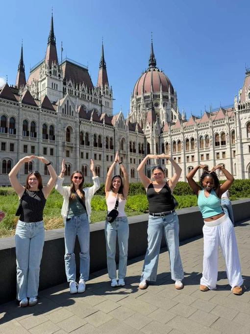 A group of people posing with arms forming letters in front of the Hungarian Parliament Building in Budapest, with a clear blue sky.