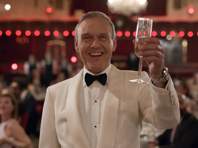 Anthony Head smiles in a white tux in the middle of a party, holding up a glass of champagne.