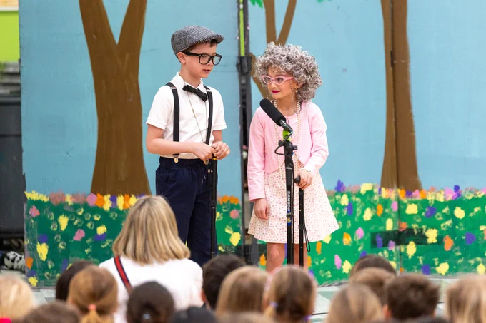 Two children dressed as elderly characters perform on stage in a school play. The scene is set with a backdrop of painted trees and flowers, creating a cheerful atmosphere.
