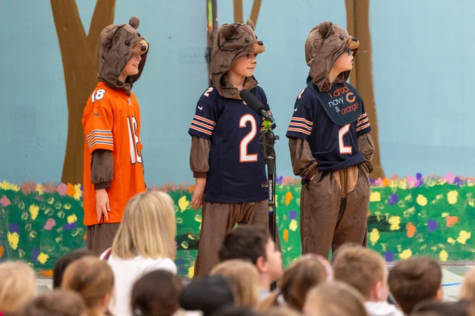 Three children dressed in bear costumes with sports jerseys are performing on stage in front of an audience. The scene is lively and playful.