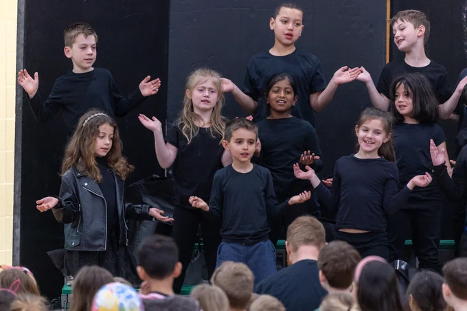A group of children on stage during a school performance, dressed in black. They appear engaged and energetic.