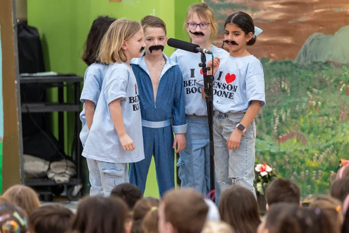 Children in fake mustaches stand on stage speaking into a microphone during a school performance. They are wearing themed shirts and one child is dressed in a glittery blue outfit.