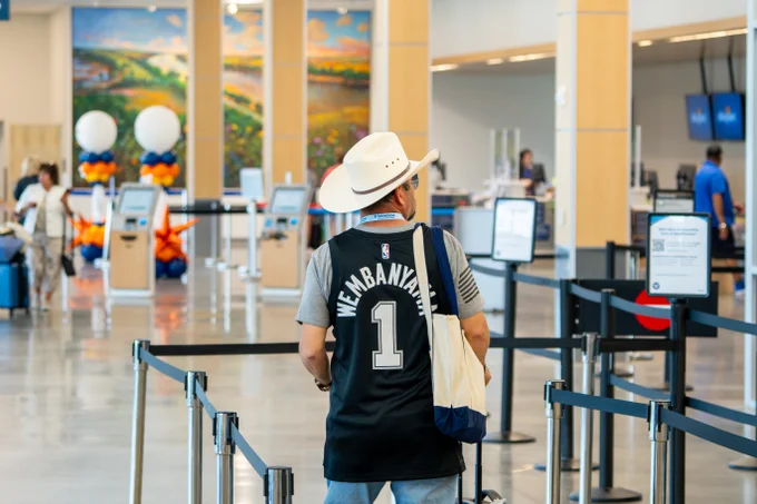 Man wearing a white cowboy hat and a black NBA jersey with 'Wembanyama 1' stands in an airport check-in line.