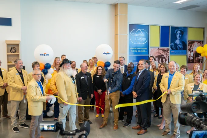 Group of people in yellow jackets and formal wear gathered for a ribbon-cutting ceremony inside a building with Allegiant balloons and Columbia College signage.