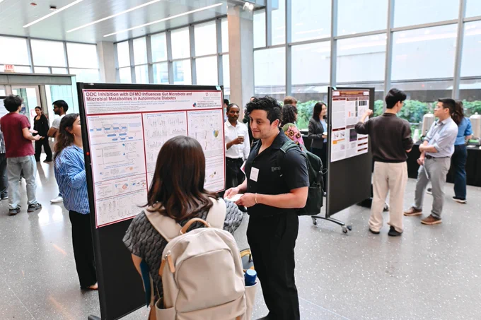 A busy indoor poster session. In the foreground, a woman views a poster titled 'ODC Inhibition with DFMO Influences Gut Microbiota and Microbial Metabolites in Autoimmune Diabetes.' A man in a black shirt, holding a plate, stands beside her. Other attendees engage with posters in the background. Large windows line the room.