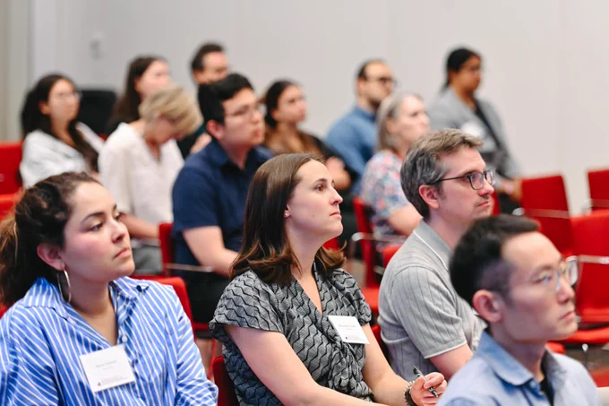 A diverse group of adults seated in red chairs, attentively looking towards a speaker during a conference or lecture. In the foreground, name tags are visible on attendees, including \"Rosemary Calabro\" on a woman in a patterned top and \"Olivia Chmura\" on a woman in a striped shirt.
