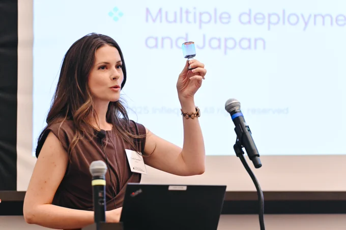 Caitlin Carnahan from Infleqtion presents, holding a small iridescent object in her raised right hand. She wears a brown top and gold bracelet. A laptop and microphones are on the table. In the background, a screen displays text: \"Multiple deployme and Japan\" and \"2025 Infleqs. All rights reserved.\"