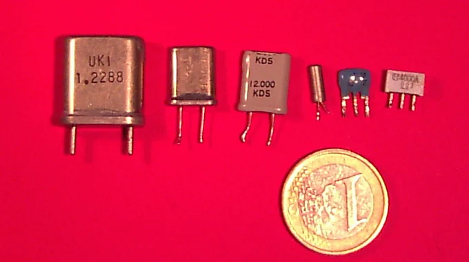 Photo of several quartz crystal resonators in different metal packages arranged on a white background, showing cylindrical and rectangular can styles with wire leads.