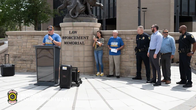 A group of people stand outside the Law Enforcement Memorial in front of a statue. One person is speaking at a podium, while others, including several in police uniforms, stand nearby. The Wichita Police Department logo is visible in the image.